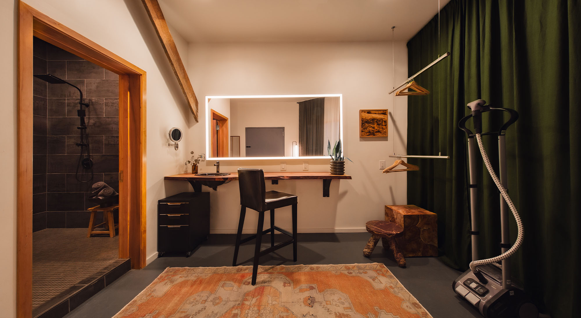 A wide photo of the greenroom at Particle Studio in Seattle, WA. There is a beautifal makeup counter surrounded by a clothing rack, steamer, and a chair. The shower in the greenroom is visable through an open door.