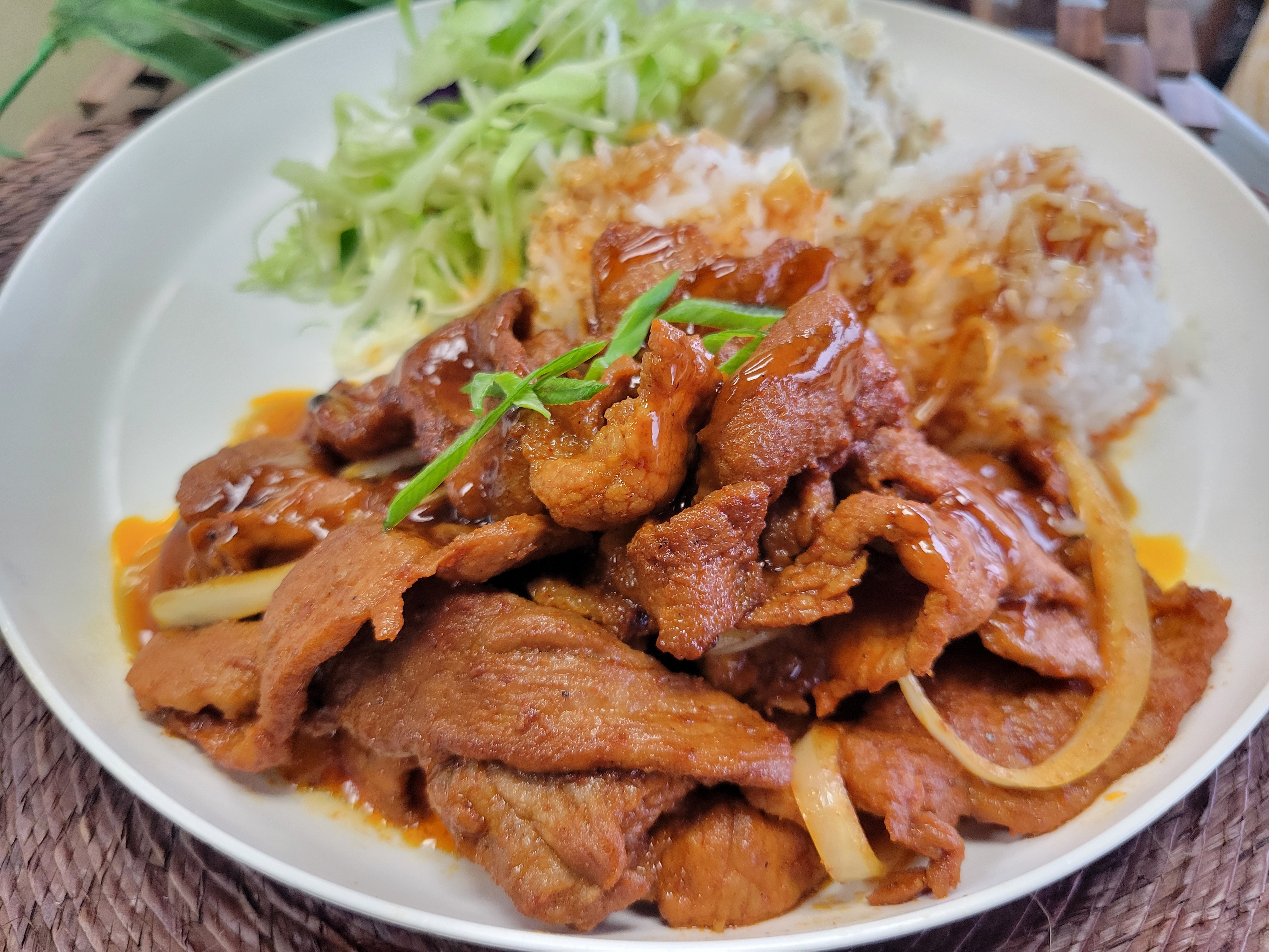 A vibrant plate of Spicy Grilled Pork wok-fried with onions, served alongside two scoops of Jasmine rice, mac salad, and a fresh Asian slaw.