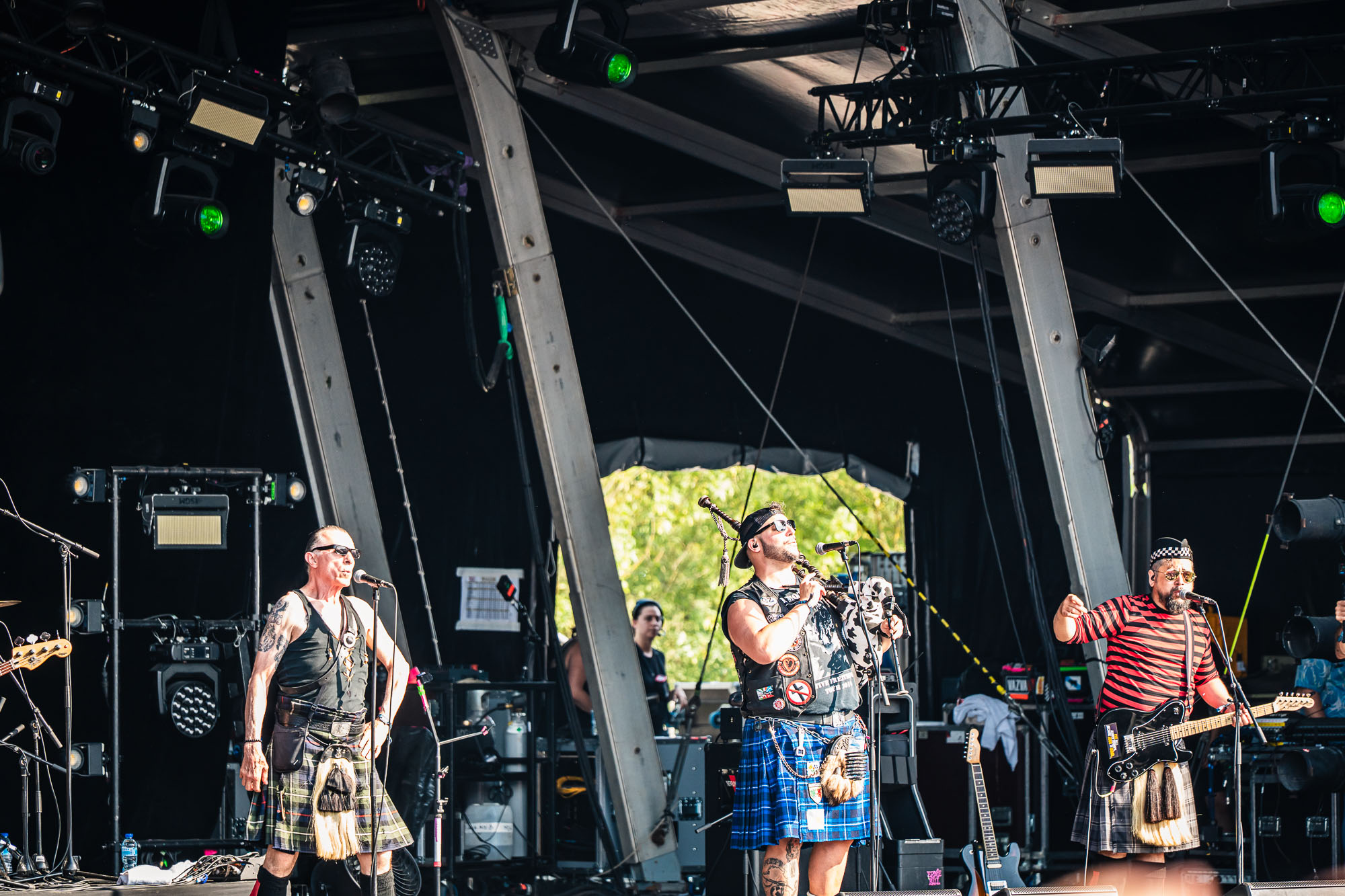 Trio de musiciens en kilt sur scène, cornemuse au centre, ambiance festive en plein jour