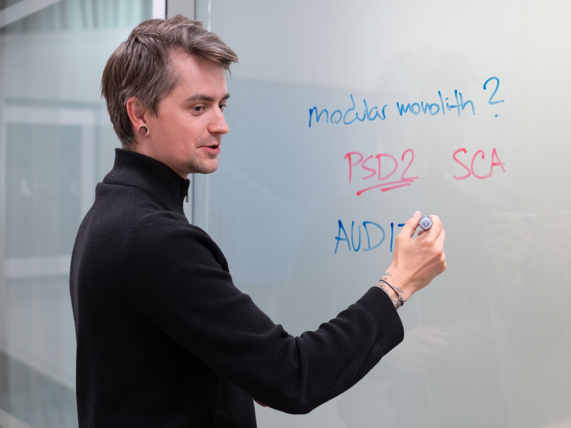 Fintech engineer writing core architecture and regulatory concepts on a glass whiteboard — modular monolith, PSD2, SCA, audit — capturing the design decisions behind building a compliant fintech platform.