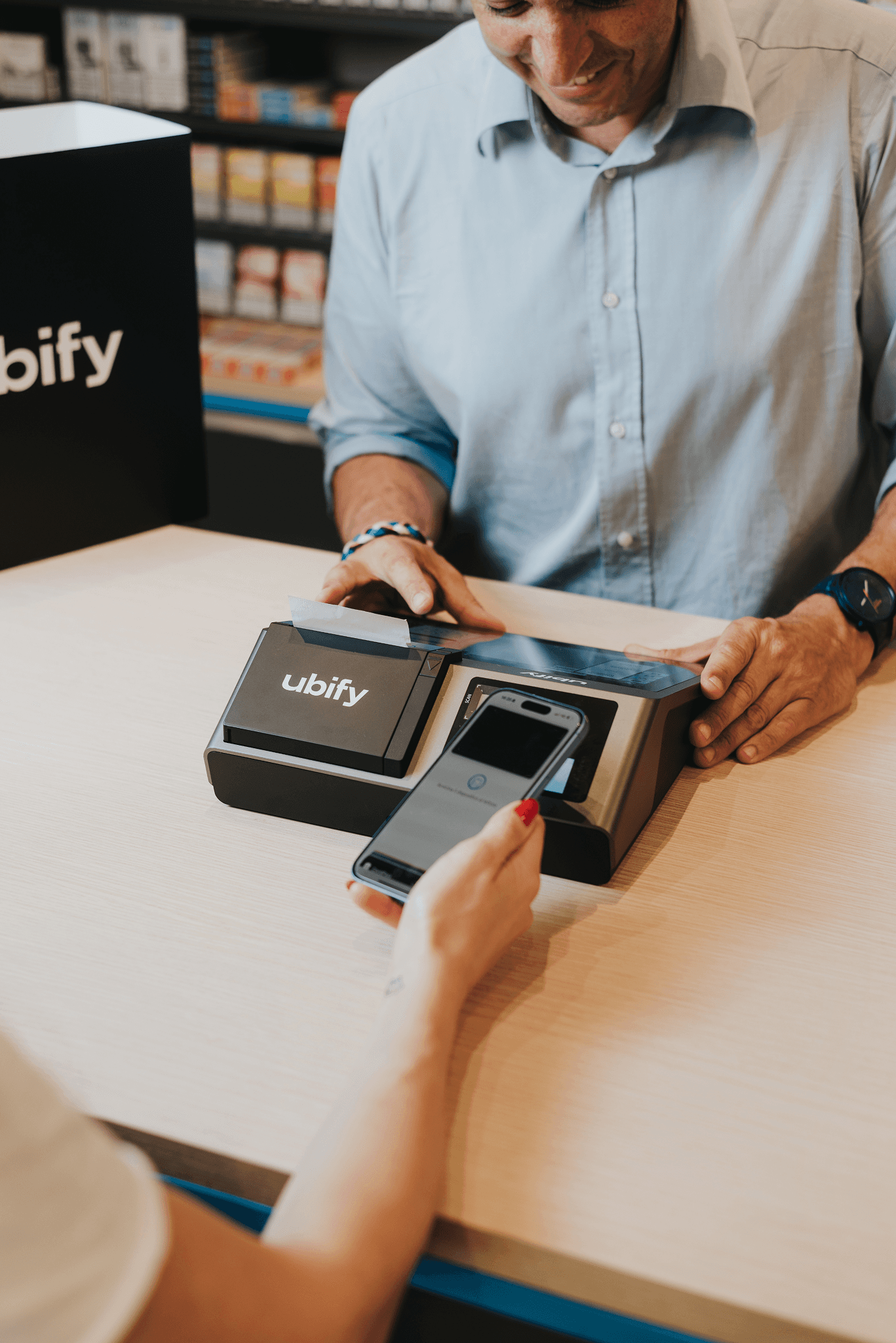 Cliente effettua un pagamento contactless con smartphone sulla Cassa Ubify, assistito da un commerciante sorridente.
