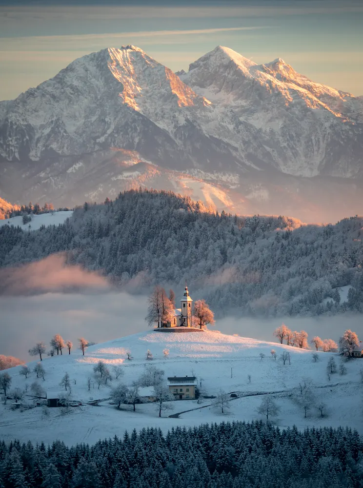 Snow-covered mountains rise majestically in the background, with a soft glow illuminating the landscape of Sveti Tomaž, Slovenia during winter morning.