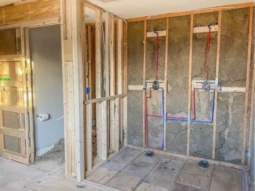 Partially constructed room with exposed wooden framing and wiring. Plumbing pipes are visible against an unfinished wall, suggesting a bathroom installation.