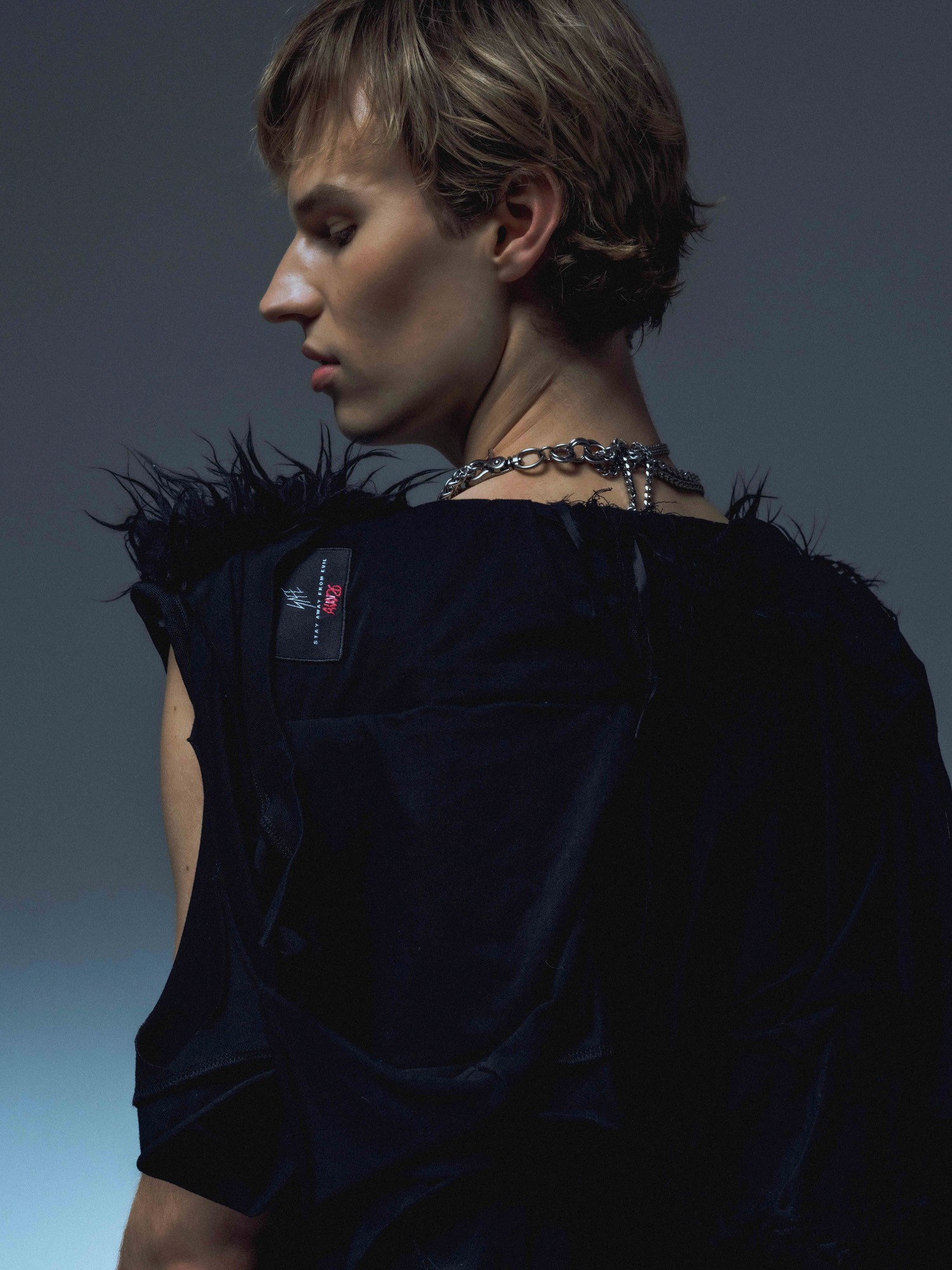 A person with short hair wears a black, feather-accented sleeveless top and a chunky silver necklace, posing against a dark background.