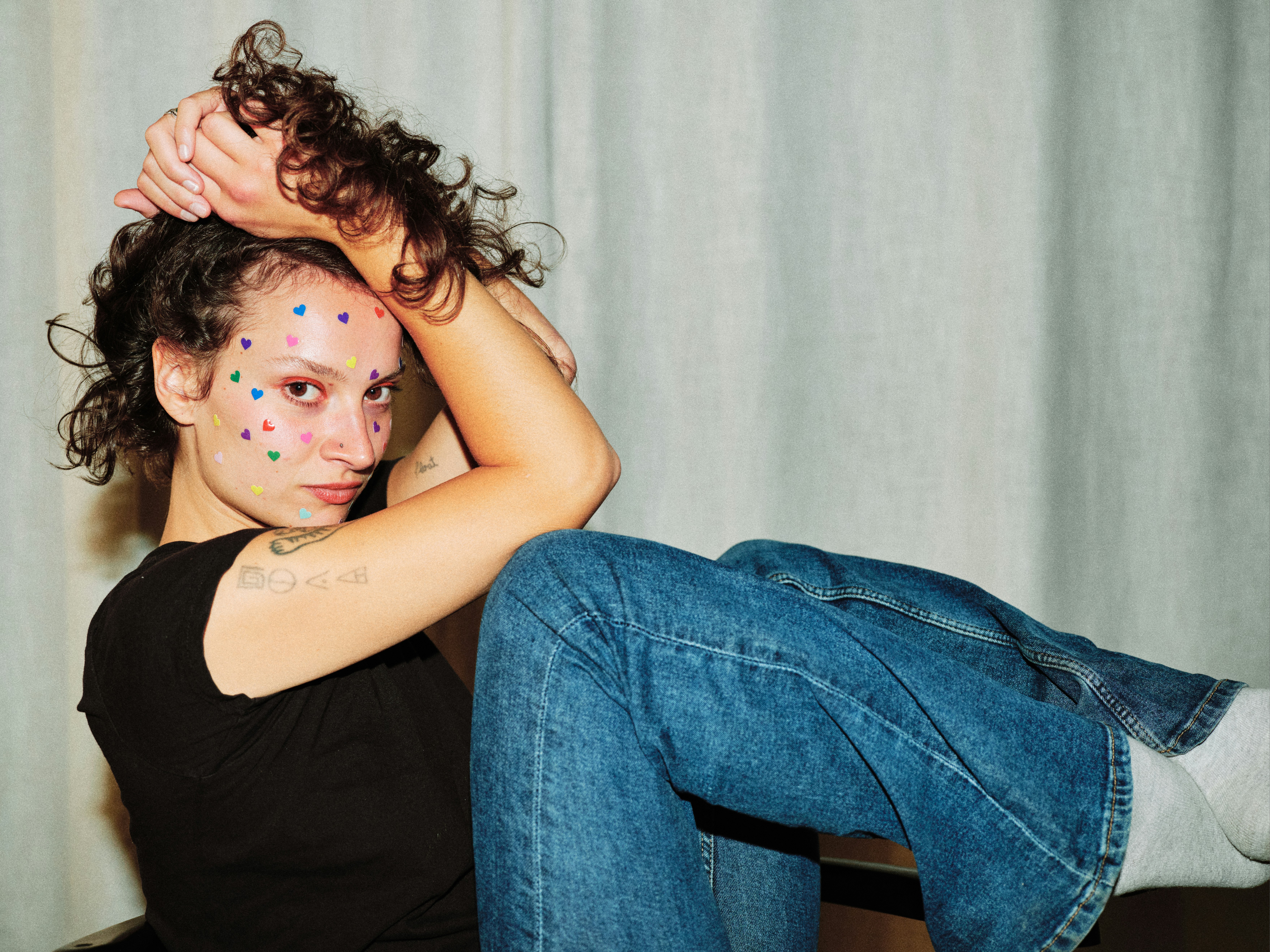 Relaxed editorial portrait of a tattooed model with multicoloured heart stickers on her face, wearing a black top and wide-leg jeans, seated against a grey curtain