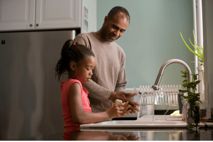 Father helping a child wash hands at a kitchen sink.