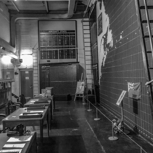 A black-and-white room with tables, ladders, large wall maps, and a board listing aircraft statuses. Flags and documents are on display.
