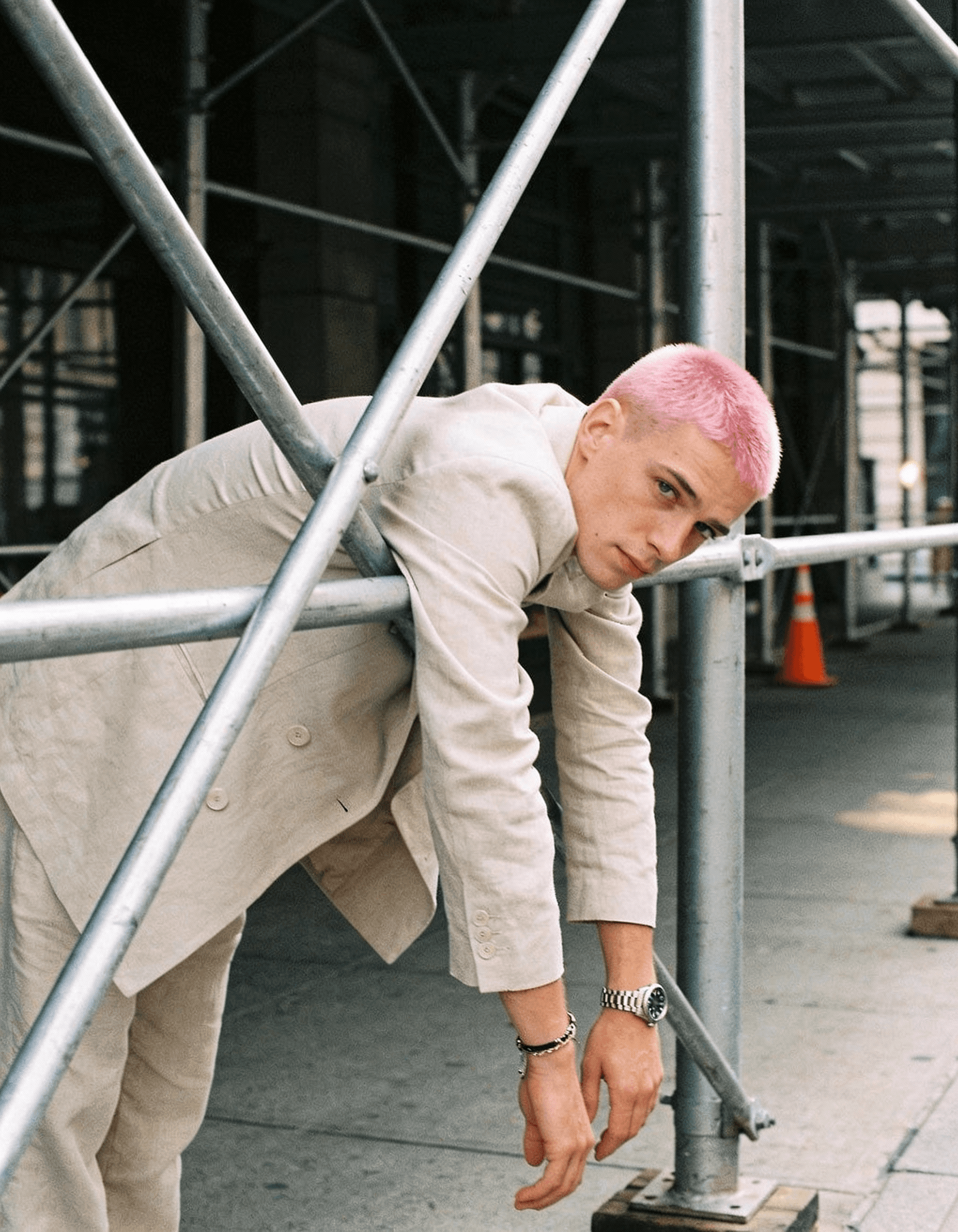 Person with pink buzz cut hair wearing a beige double-breasted blazer and gray pants, leaning against metal scaffolding railings under an industrial structure with a silver watch and bracelet visible