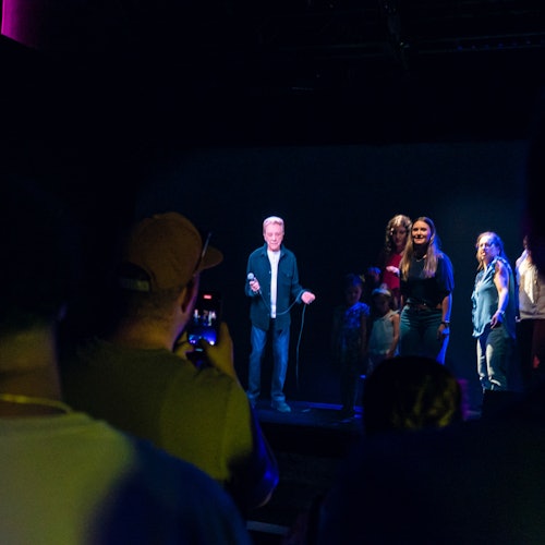 一群人站在灯光昏暗的舞台上，而坐在观众席上的人则观看并拍照。