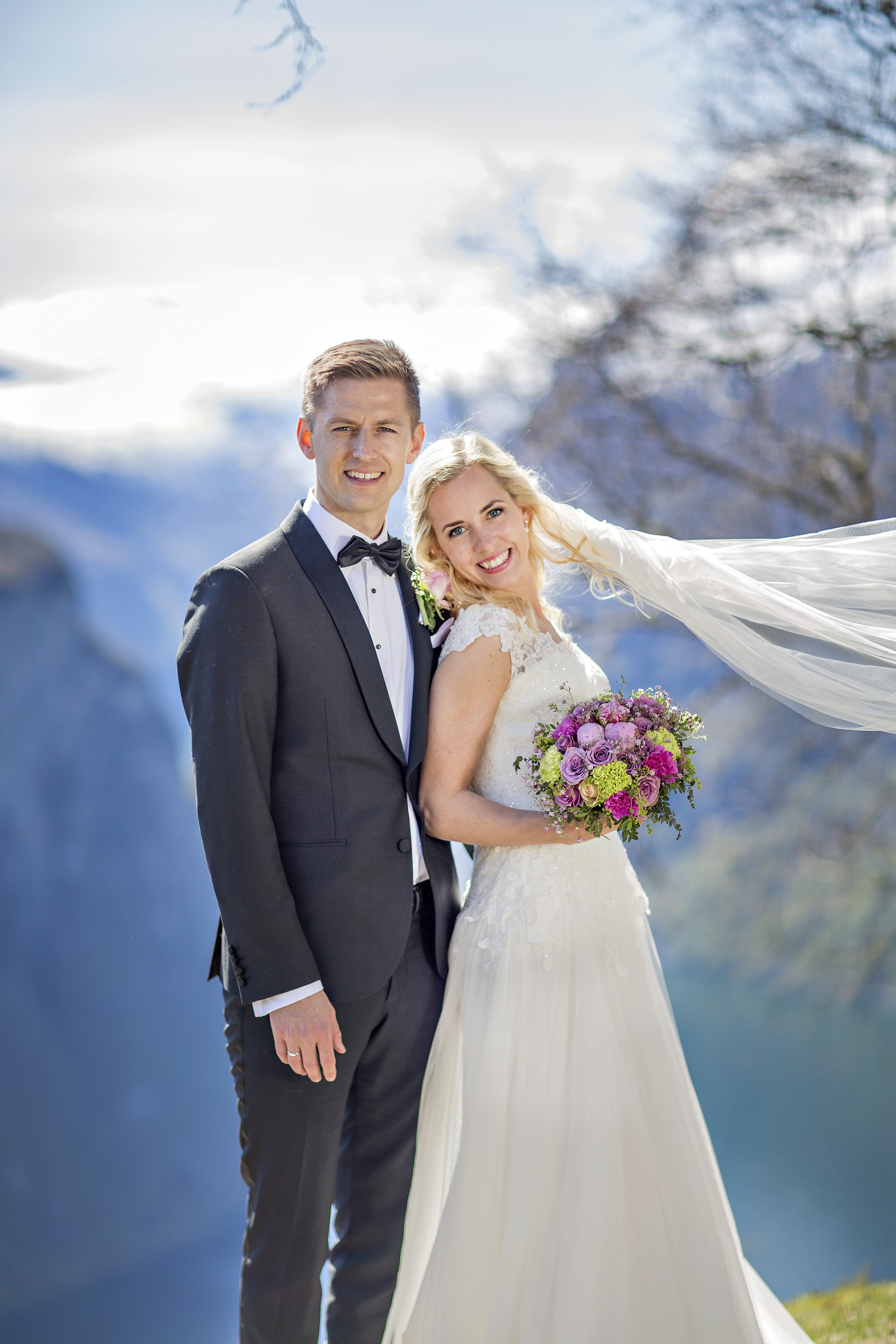 A bride and groom in formal attire stand joyfully under sunlit autumn trees on a cliff edge, overlooking a scenic mountain valley.