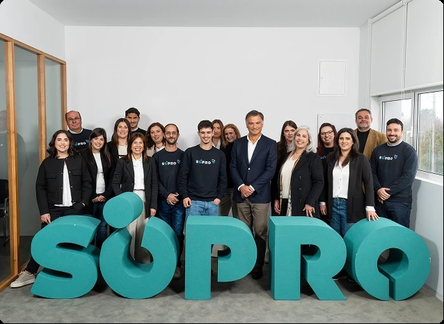 Grupo de pessoas sorrindo ao lado de grandes letras 3D formando a palavra  SRO, em um ambiente corporativo moderno, destacando a equipe da S/PRO na campanha de destaque na área de tecnologia e inovação.