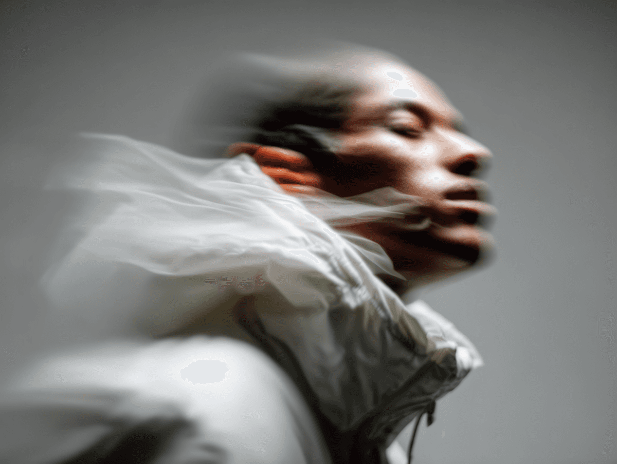 A blurred portrait of a person in a white jacket, showcasing motion and energy with a neutral background.