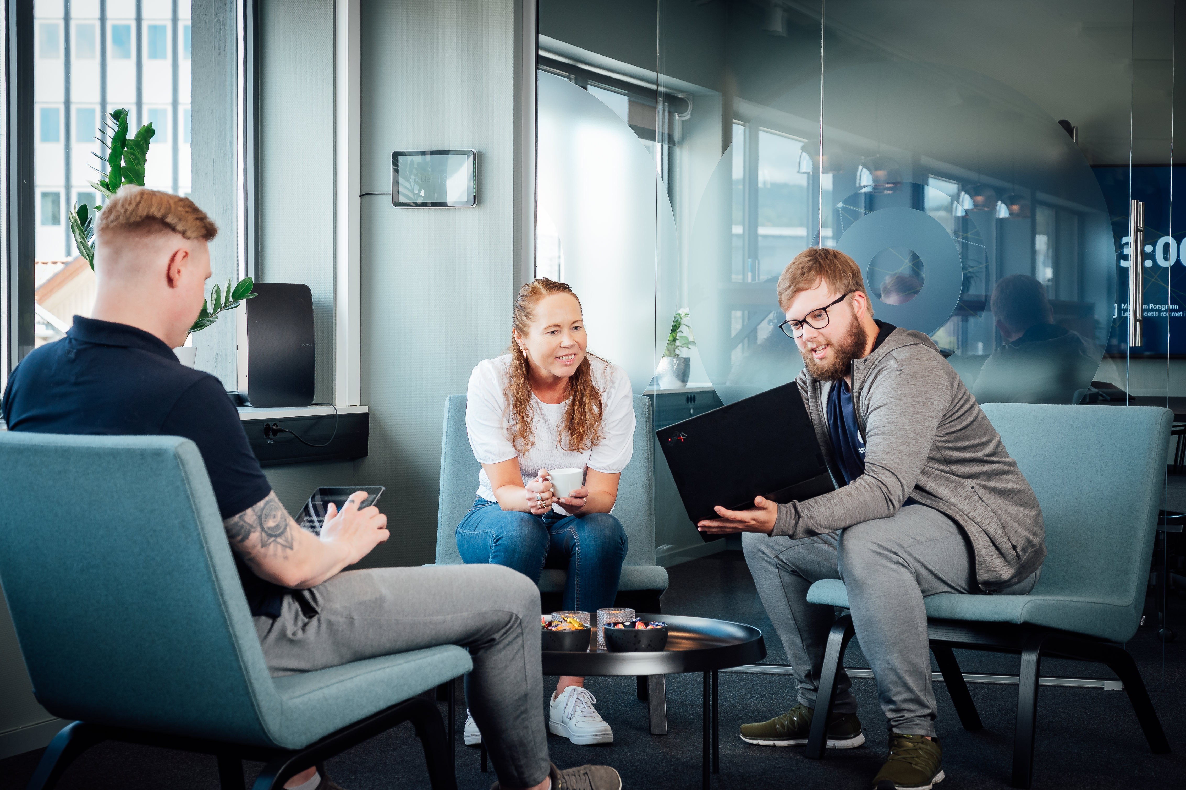 Tre kolleger sitter i en uformell møte­sone og diskuterer rundt et lite bord, med PC og notater i et moderne kontormiljø.