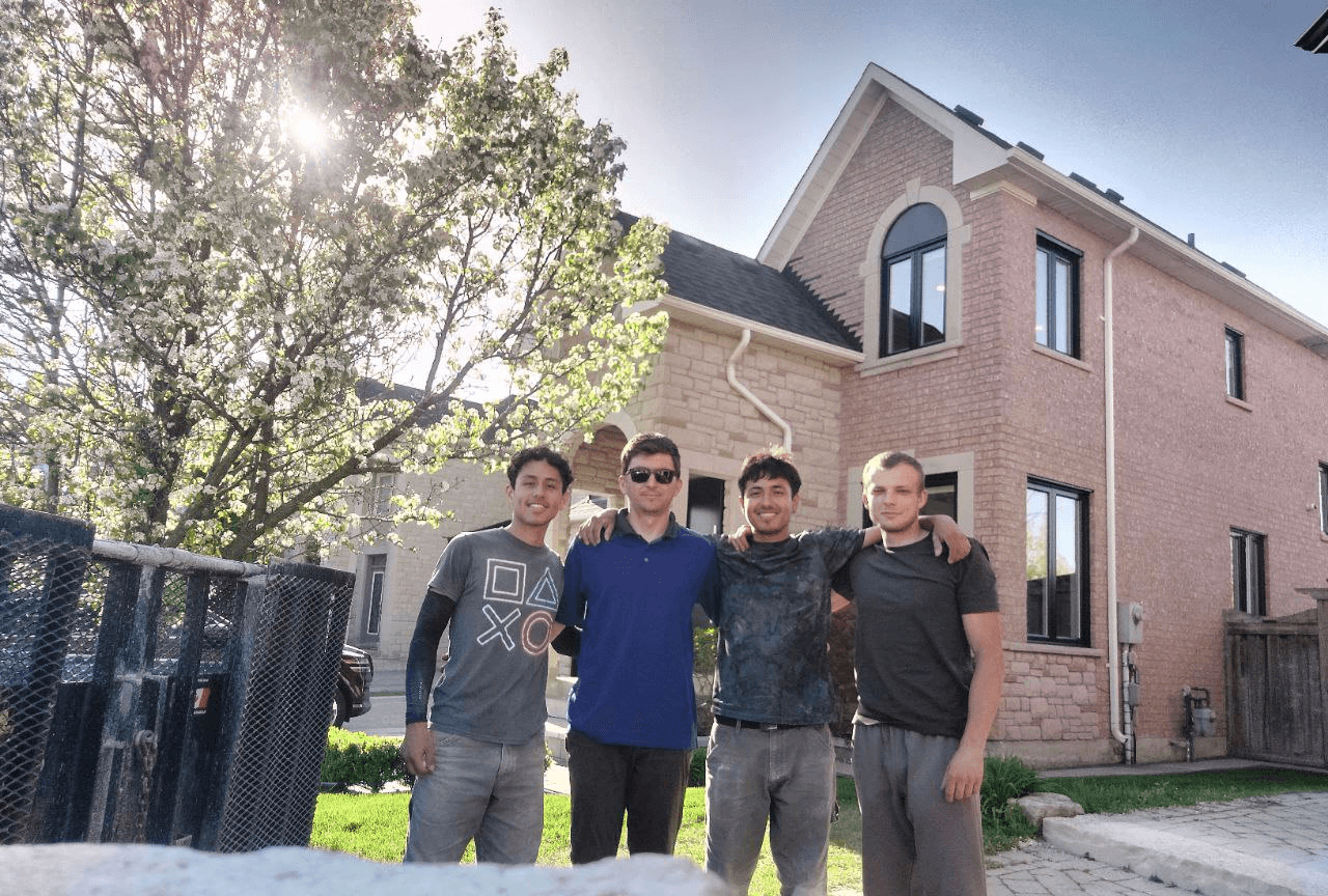 Four individuals pose together outside a pink house with a tree in the foreground on a sunny day.