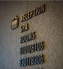 Signalétique Interne « Panneau indicateur mural avec une icône représentant un château et les mentions suivantes : « Réception », « Spa », « Chambres », suivies des numéros de chambre « B101/B106 », « B301/B306 ». »