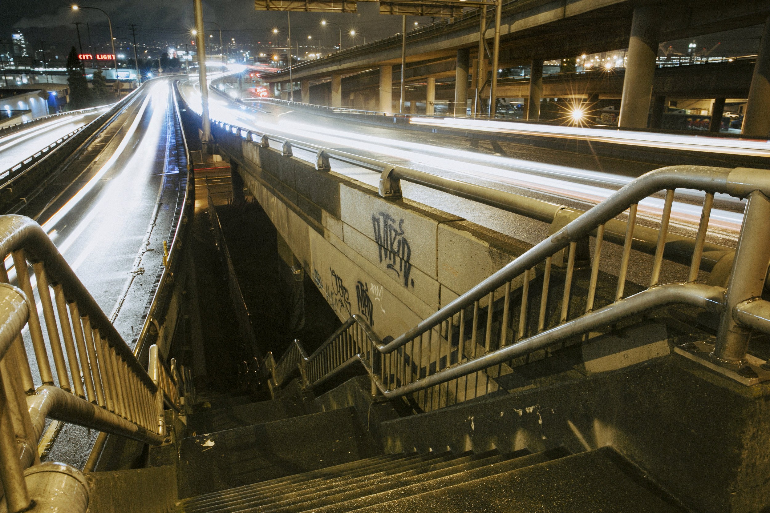 Long-exposure shot of light trails on Seattle’s SODO highway at night, with graffiti-covered stairway and illuminated overpasses.