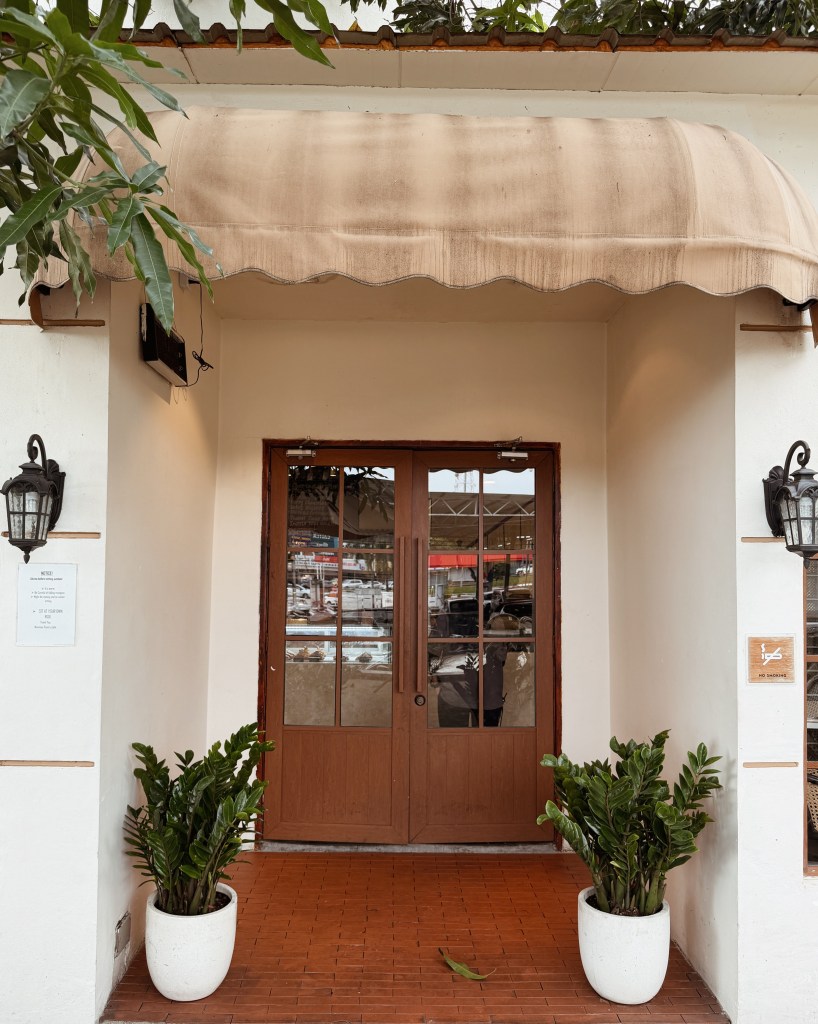 Entrance of a building featuring double wooden doors, a beige awning above, and potted plants on either side.