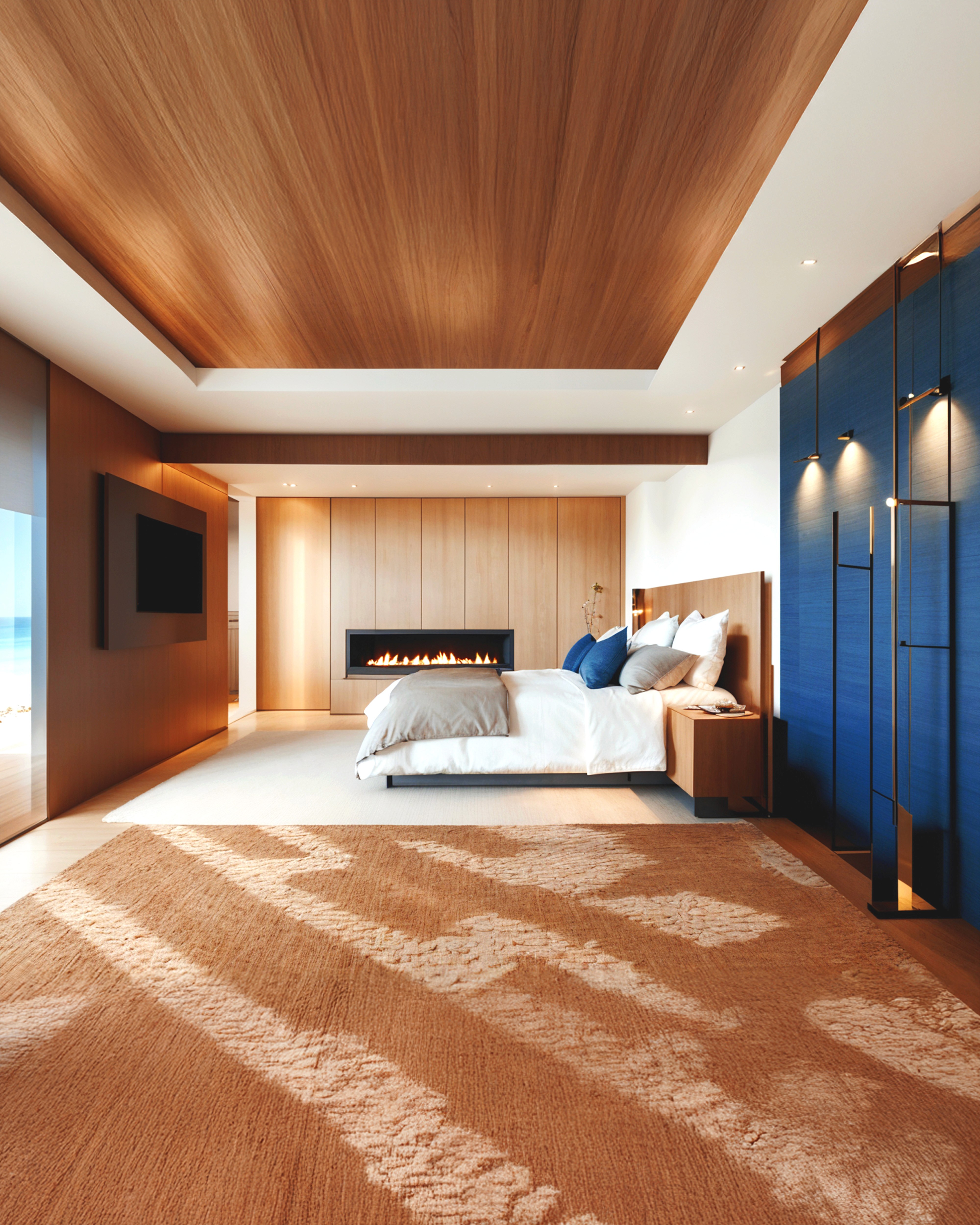 A serene bedroom featuring a wood-paneled ceiling, modern fireplace, soft textiles, and warm natural light with ocean views.