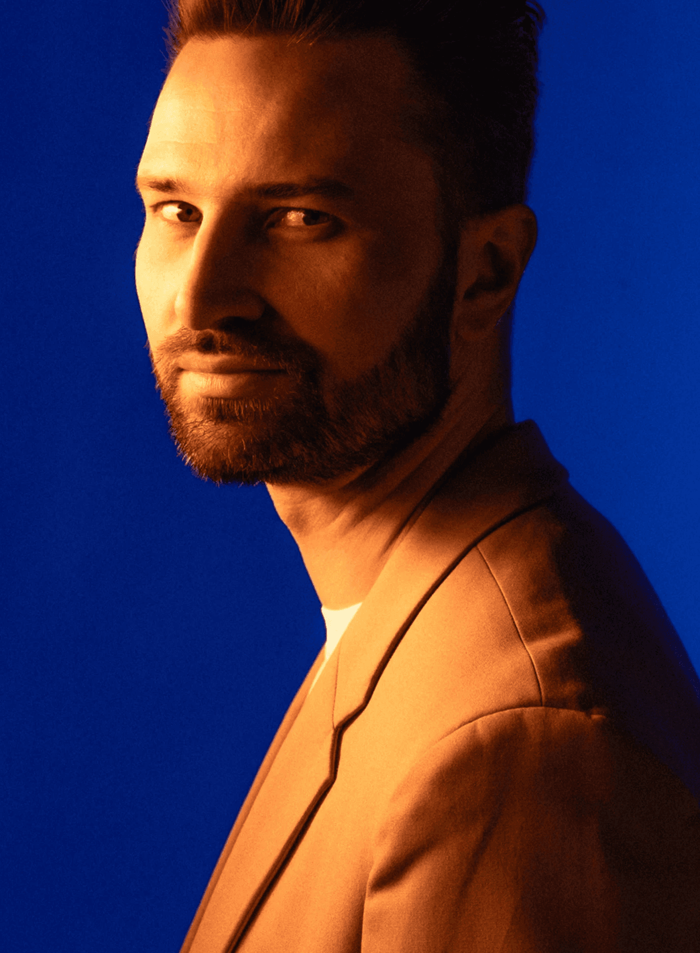 Profil boczny męskiego członka zespołu Music Shapes w ramach projektu Electric Shapes, z brodą i w beżowym garniturze, uchwycony w intensywnym, teatralnym oświetleniu w tonacji bursztynowej na niebieskim tle.