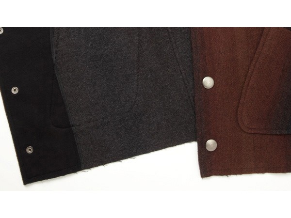 A close-up view shows the front of a brown jacket with dark gray and black panels. The left panel is black with silver rivets, while the right panel is solid dark brown with two silver rivets. The jacket appears to be made of a textured material, possibly wool or a wool blend. The image focuses on the jacket’s front closure and panel details.