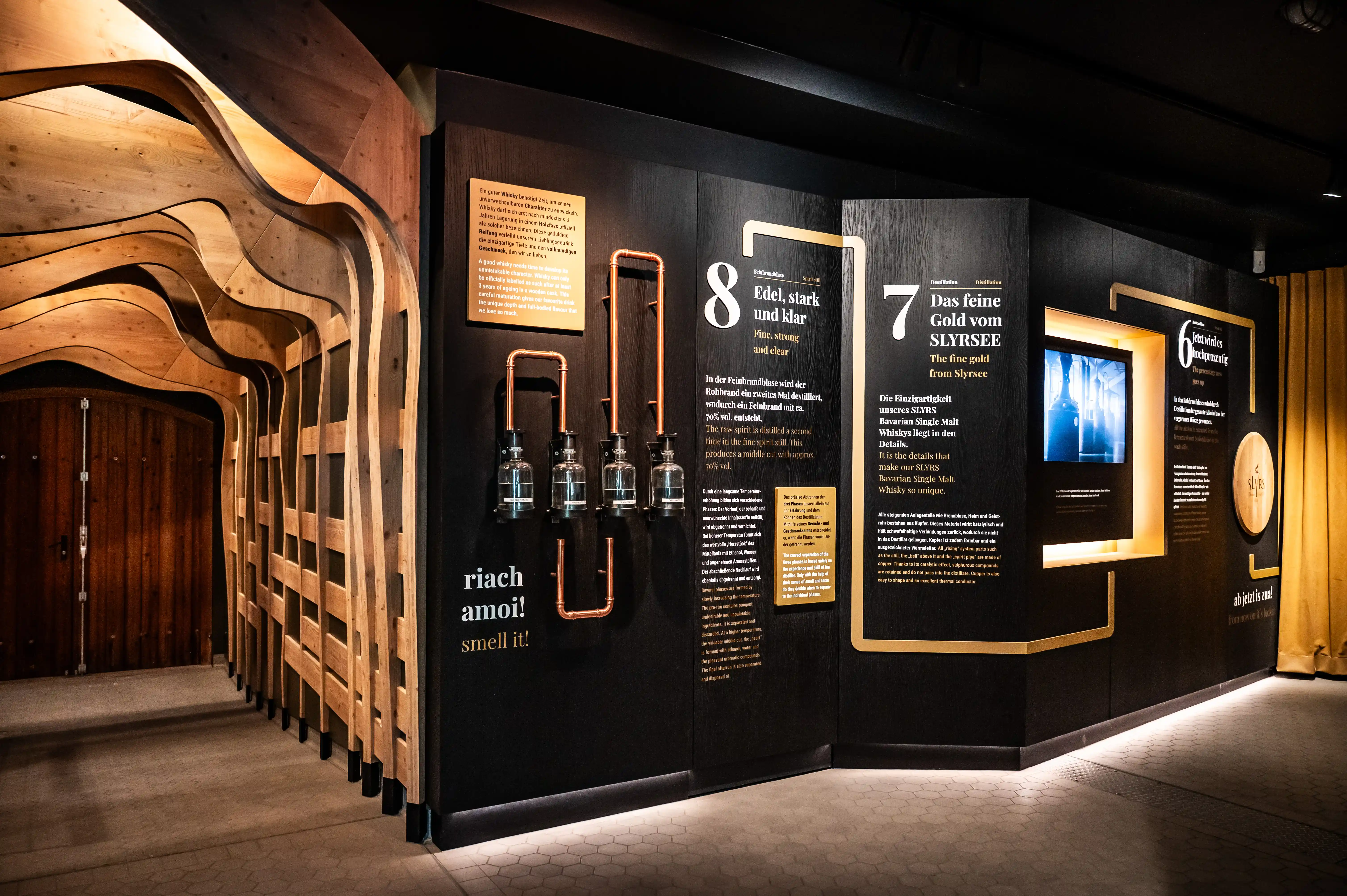 Smell-it station with flasks and copper piping along the dark wall next to the wooden tunnel at the SLYRS exhibition.