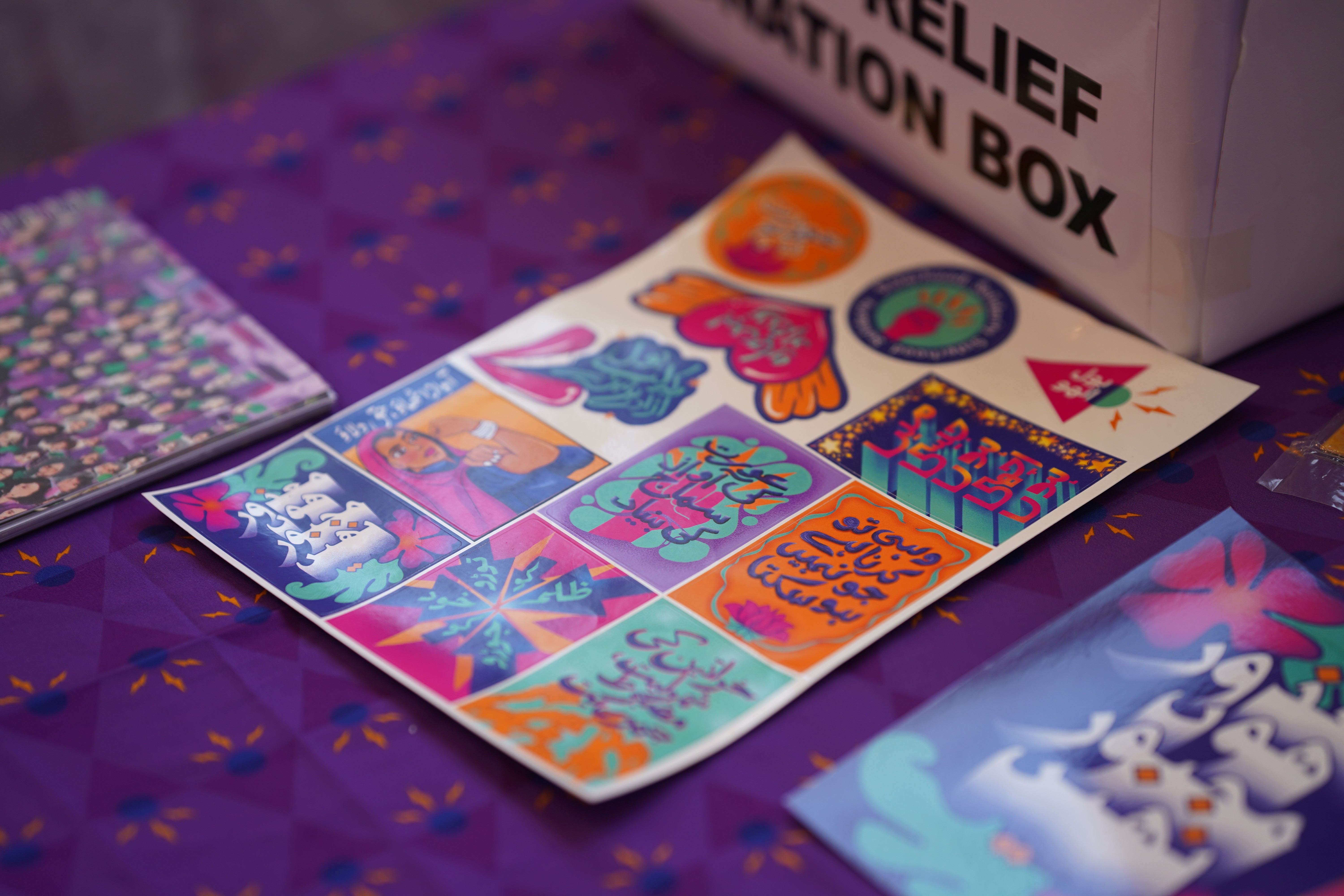 3/4 angle view of a sticker page filled with an assortment of vibrant graphic designs against a decorated purple table cloth, with more posters surrounding the sticker page
