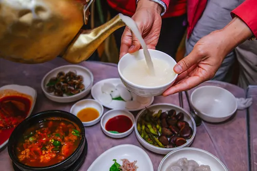 A person pours Korean rice wine (makgeolli) from a copper pot above a table with food.