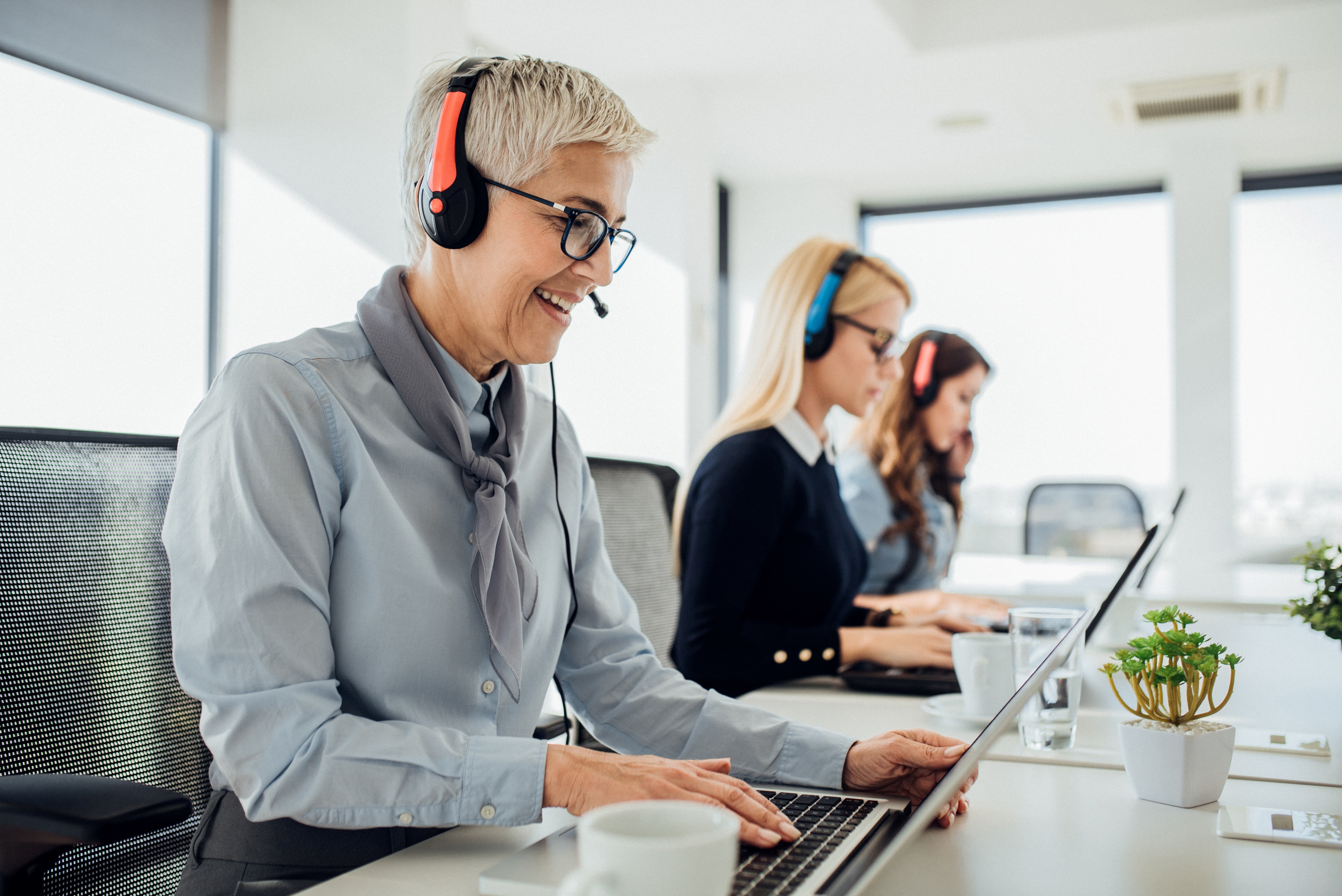 Three customer service representatives wearing headsets sit in a bright, modern office while working on laptops. They are smiling and focused, representing a professional AI-powered support team.