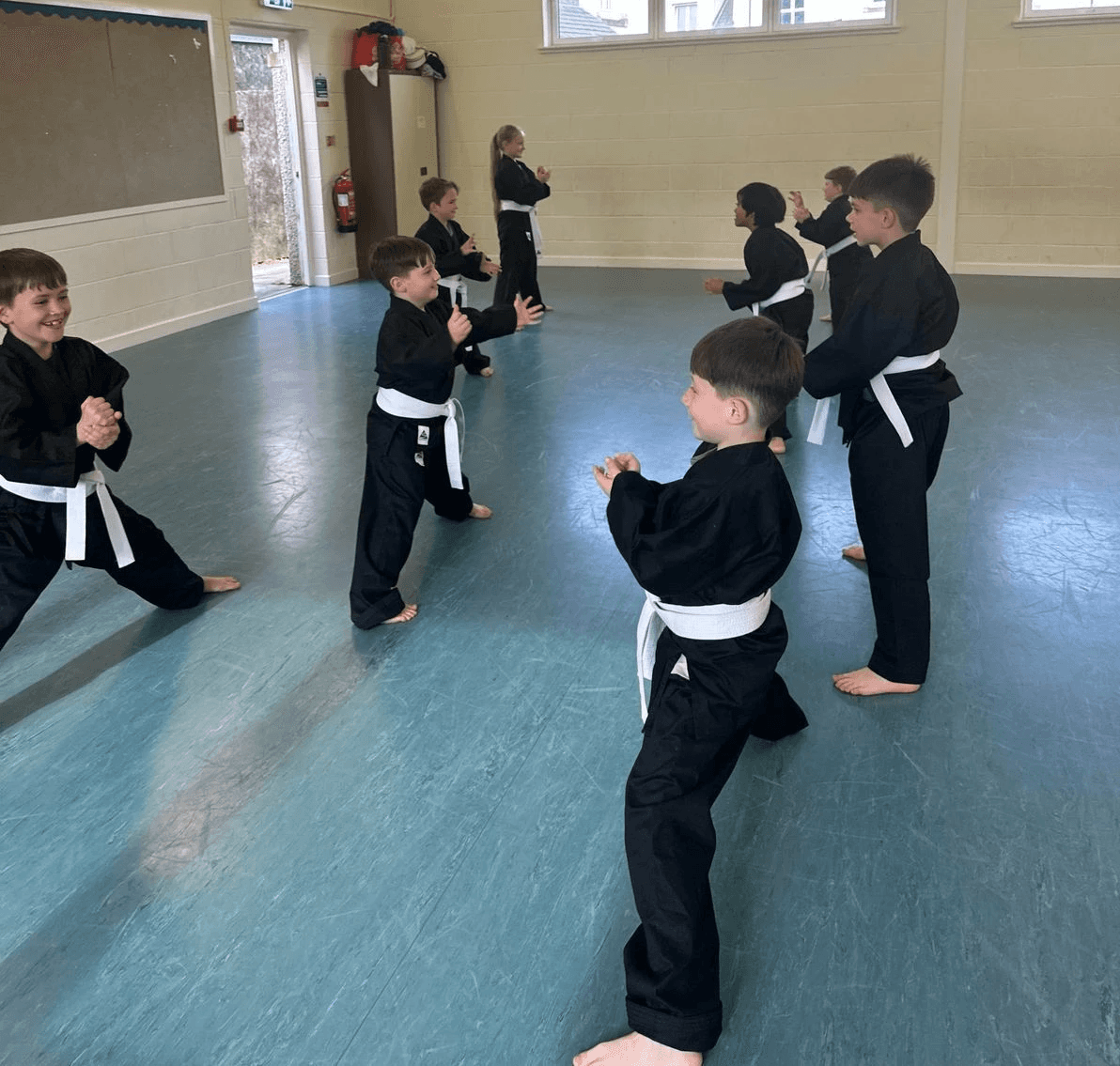 Children in black martial arts uniforms practice in a spacious hall with blue flooring. They appear focused, wearing white belts, in a light-filled room.