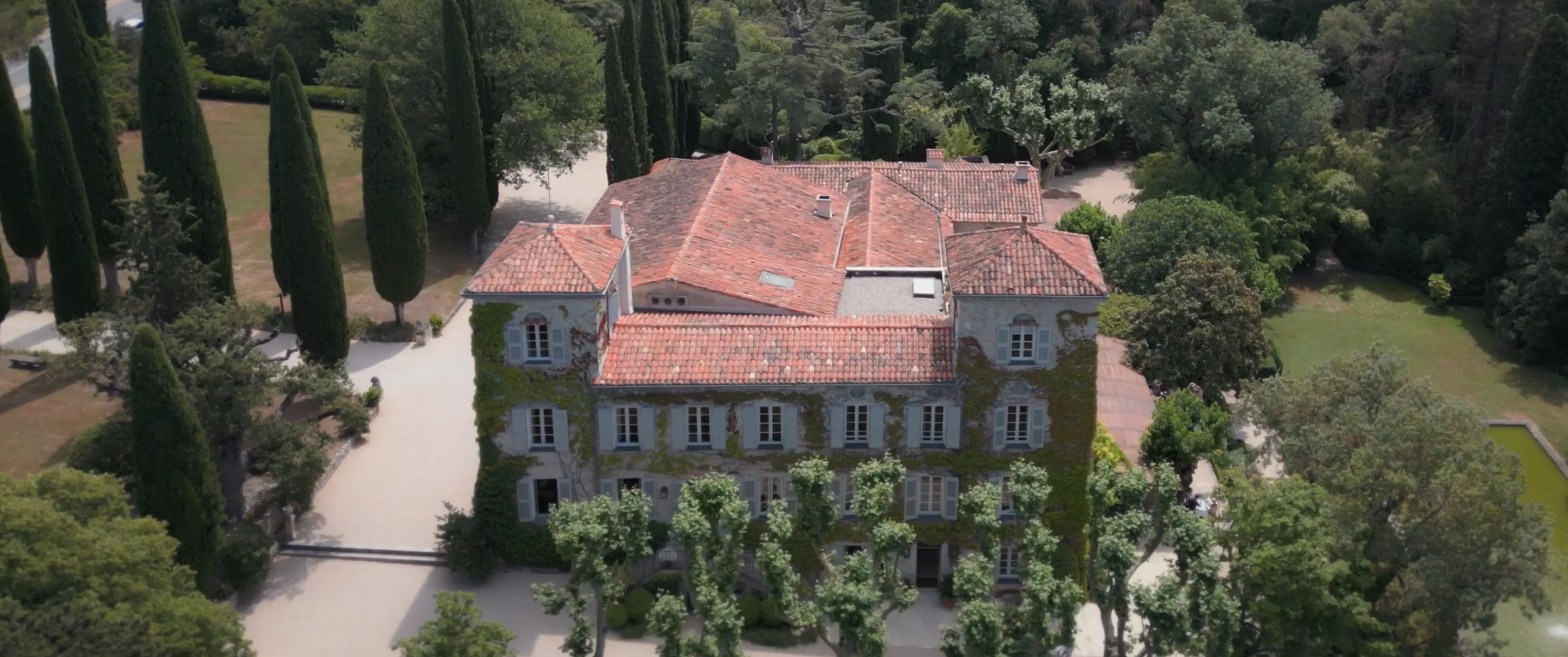 Aerial view of a picturesque stone mansion with ivy-covered walls and a terra-cotta tile roof, surrounded by lush greenery and tall cypress trees, showcasing elegant architecture and serene natural beauty.