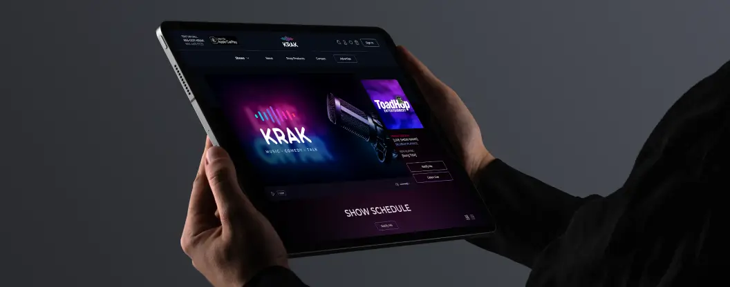 Hands holding a tablet displaying the full Krak website, featuring a neon-lit design and media show schedule.