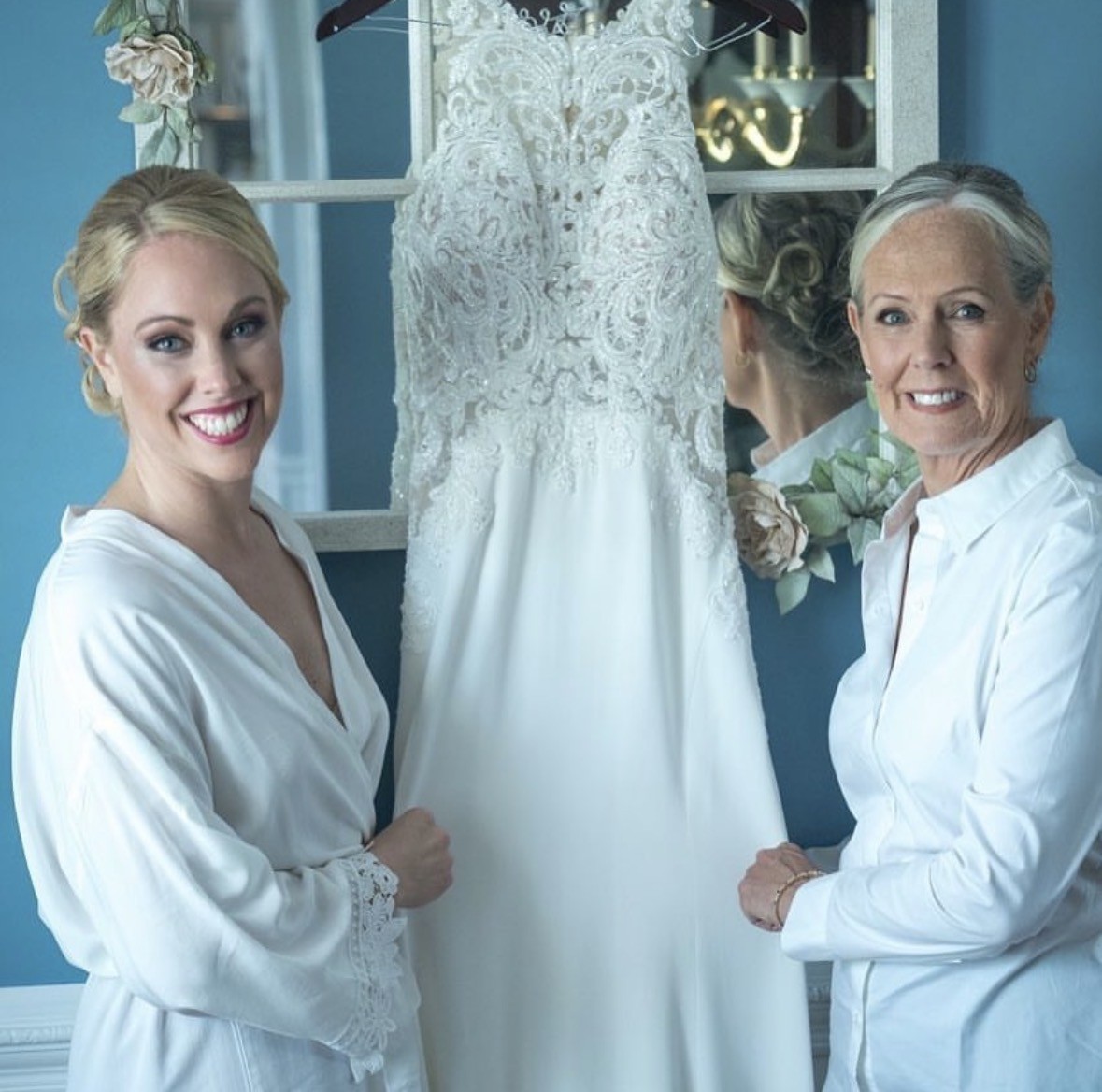 Bride and a family member preparing together beside a wedding gown against a blue wall