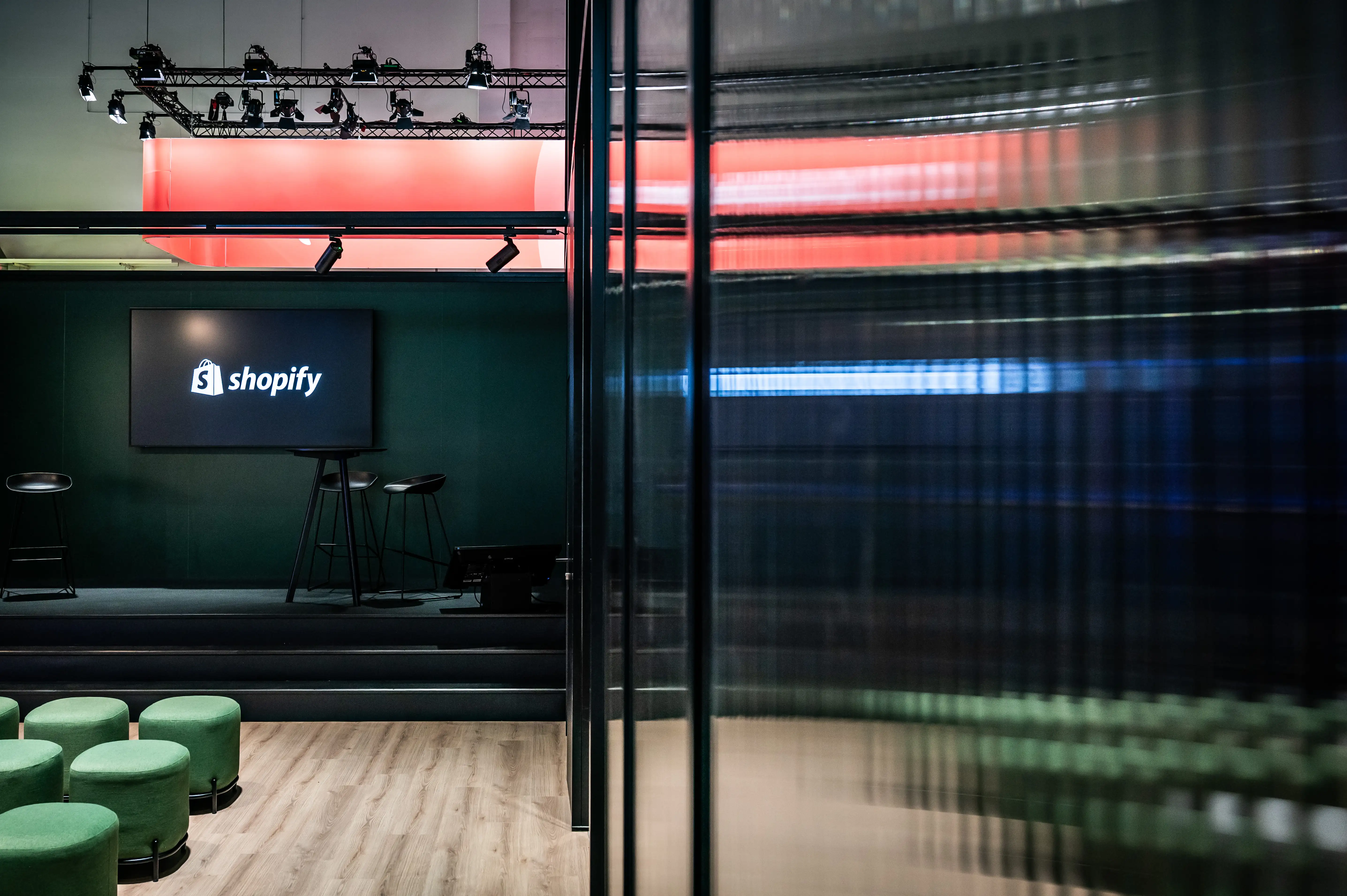 Stage area at the Shopify booth with logo screen, bar stools and green upholstered stools behind ribbed glass.