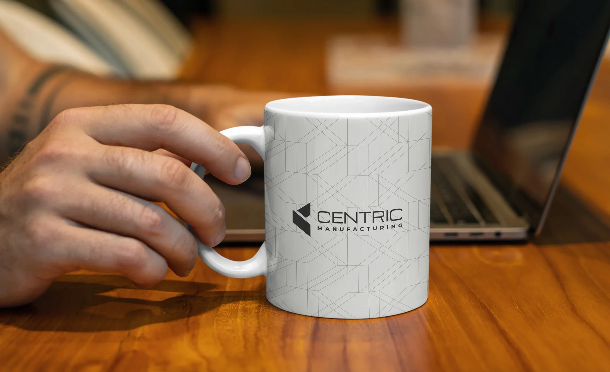 Hand holding a branded Centric Manufacturing coffee mug beside a laptop.
