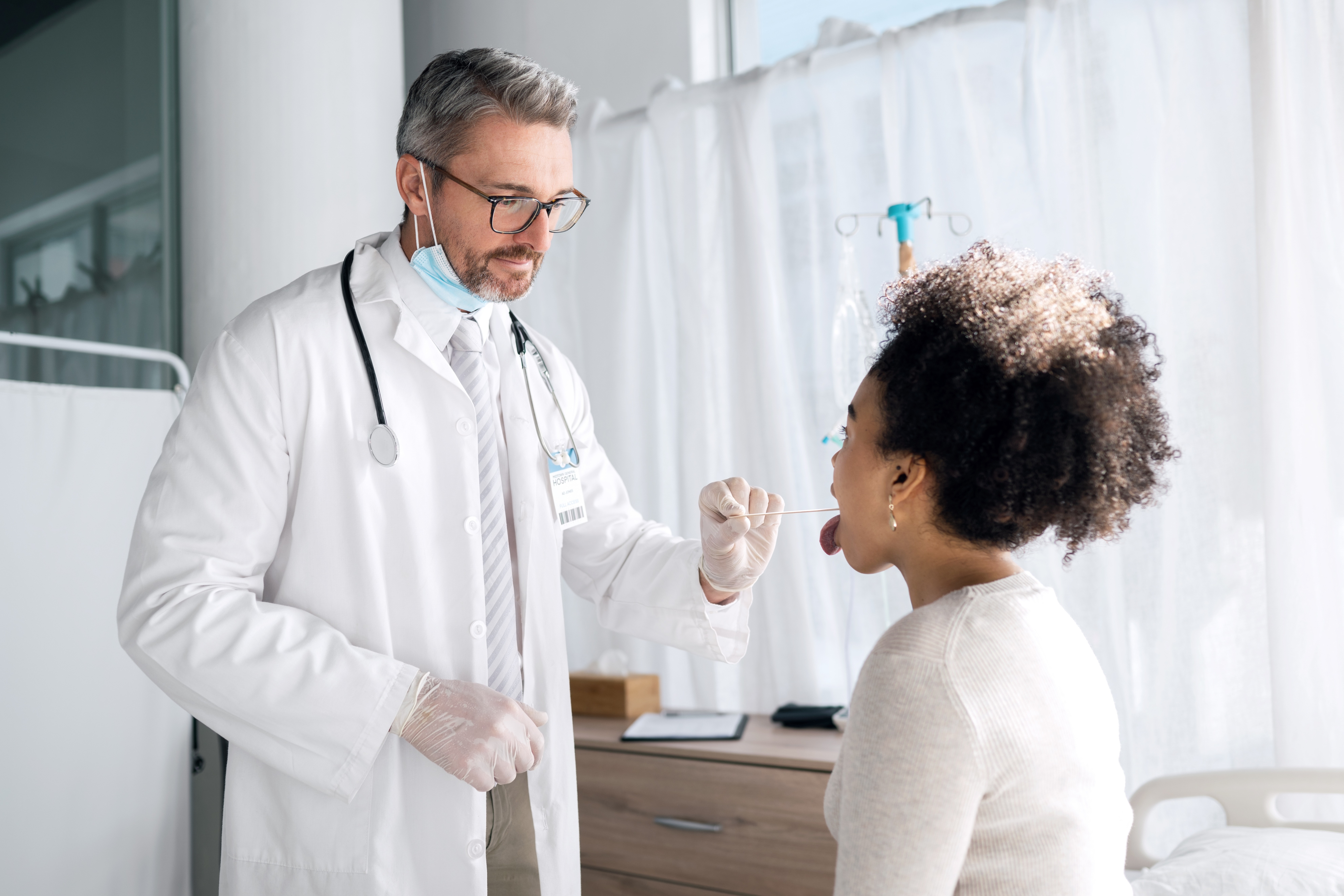 Doctor in a white coat and gloves taking a throat swab from a seated female patient in a hospital room