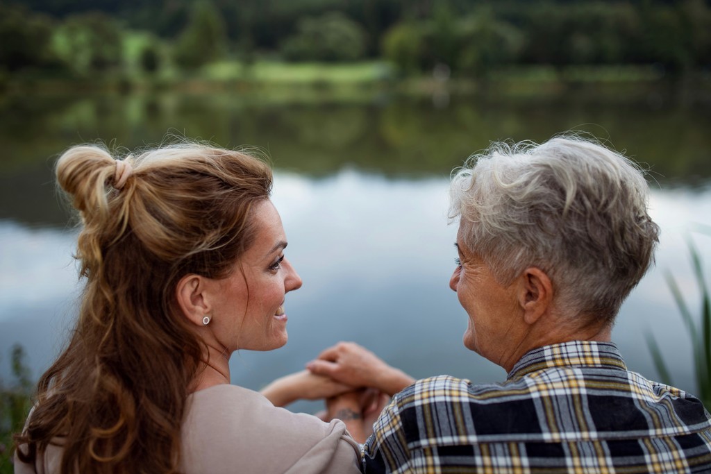 A woman and her elderly mother share a tender moment by a serene lake, enjoying the tranquil natural scenery and each other's company.