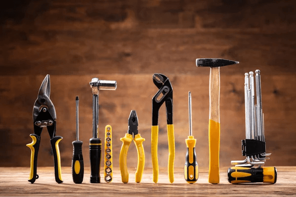 A neatly arranged set of nine tools on a wooden surface against a brown background. Tools include pliers, a hammer, screwdrivers, and wrenches, all with yellow accents.