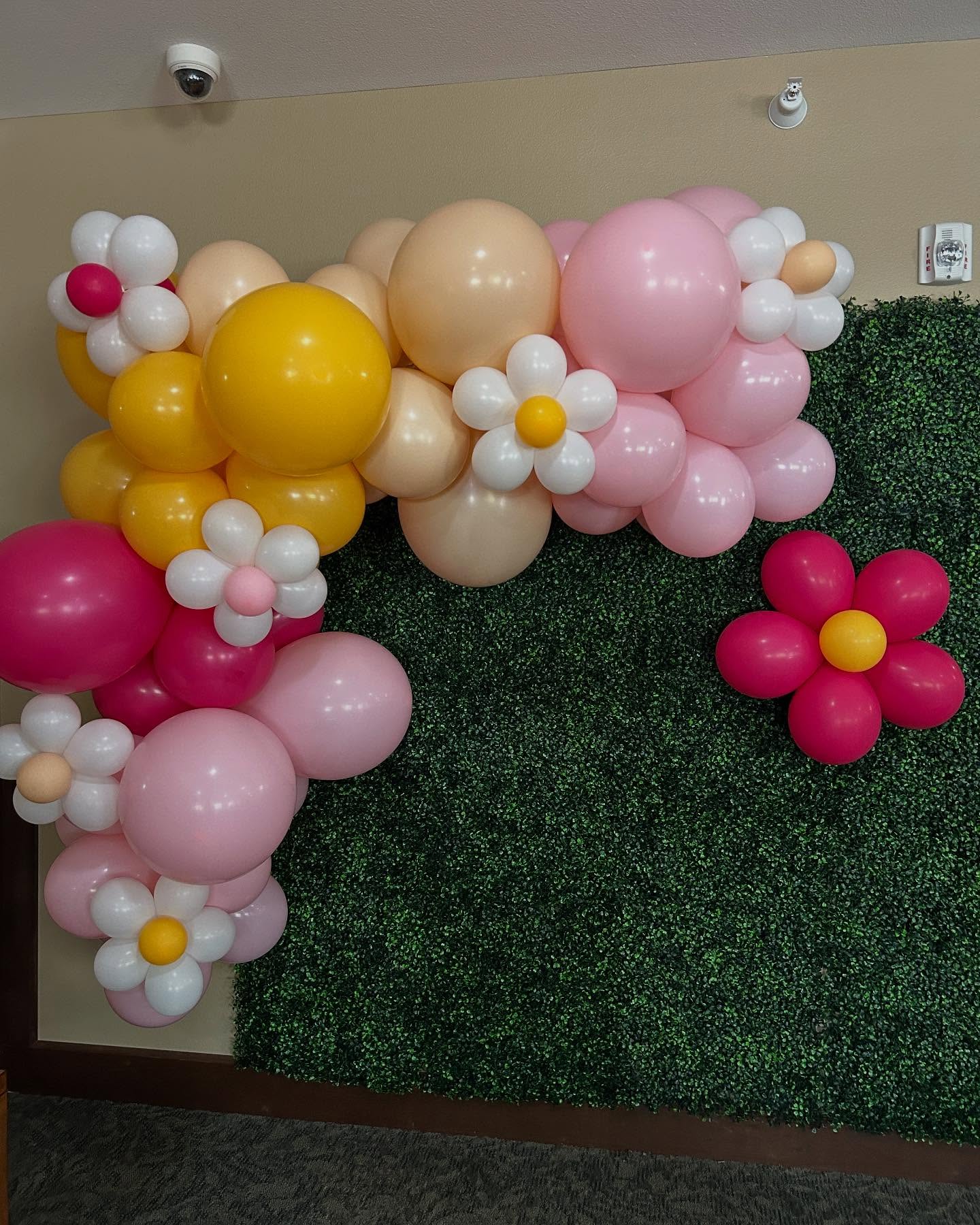 Pink, peach, and white balloon garland with flower accents attached to a green boxwood backdrop for a photo display by Be Bold Balloons