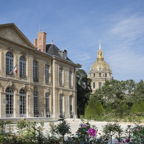 Povijesna dvokatnica s francuskom zastavom, uređenim vrtom i rascvjetanim cvijećem, dok se u pozadini vidi kupola sa zlatnim šiljkom.