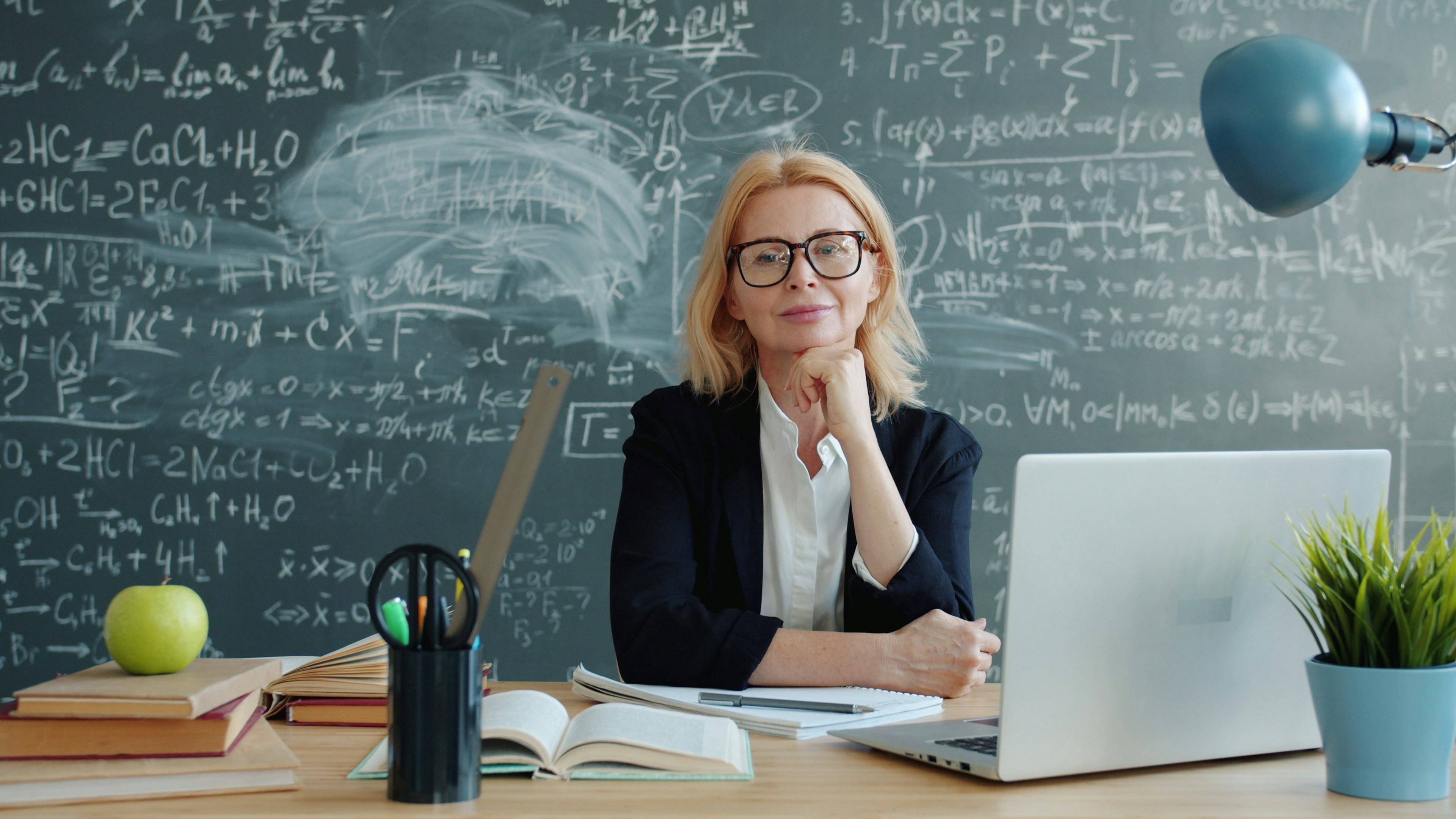 Using AI in Teaching shown by a teacher seated at a desk with books and a laptop in front of a chalkboard full of equations.