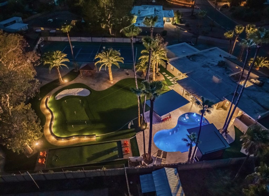 A nighttime aerial view showcases a luxurious vacation rental home featuring a unique, kidney-shaped swimming pool, a well-lit mini-golf course, and lush palm trees, offering an affordable yet upscale getaway experience.