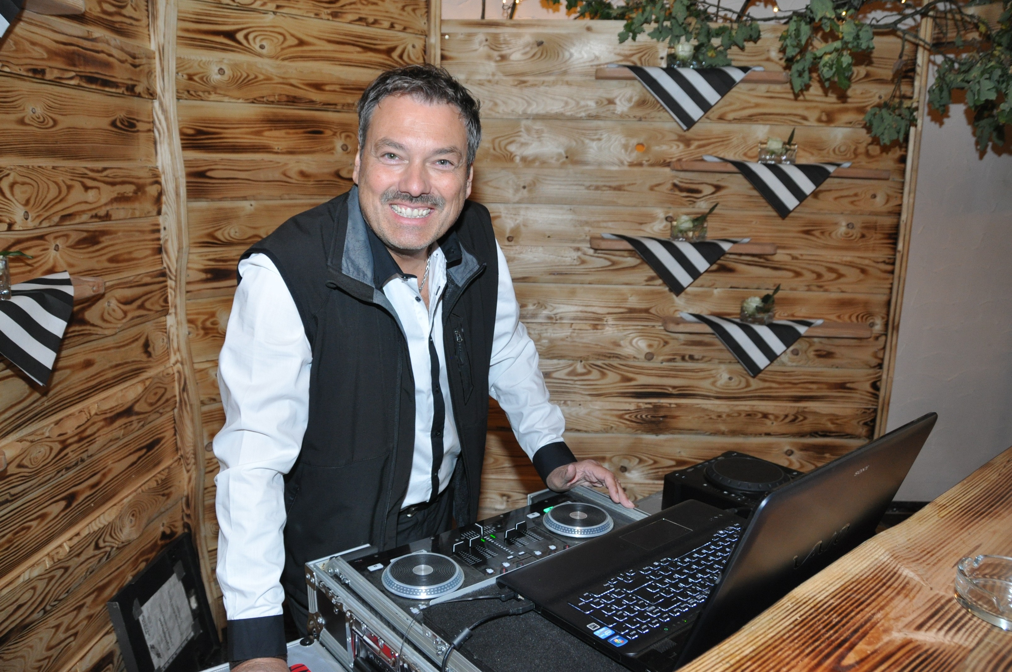 Ein Mann, freundlich lächelnd, vor DJ Equipment, beratende und offene Haltung.