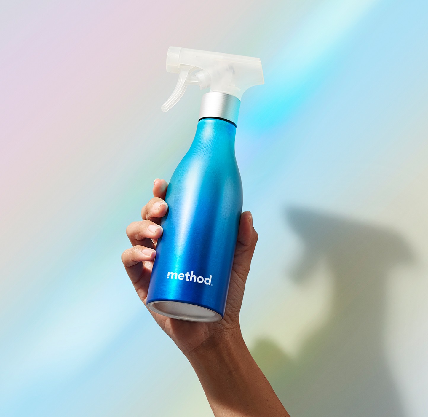Hand holding up a blue aluminum spray bottle with a clear trigger against a pale rainbow backdrop