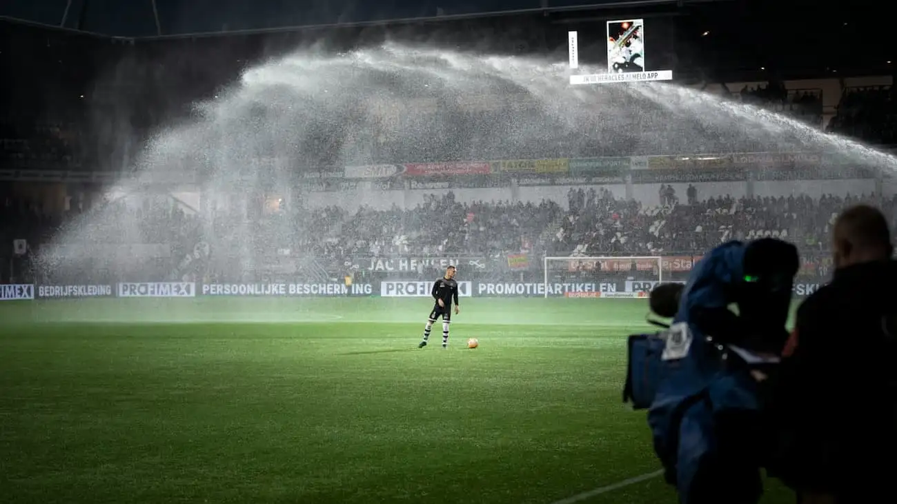 The photo shows effective pitch watering in a football stadium during preparations for an upcoming match, with a focussed player in the background. Ideal to showcase professional match preparation and the role of sponsors in providing top infrastructure for football clubs.