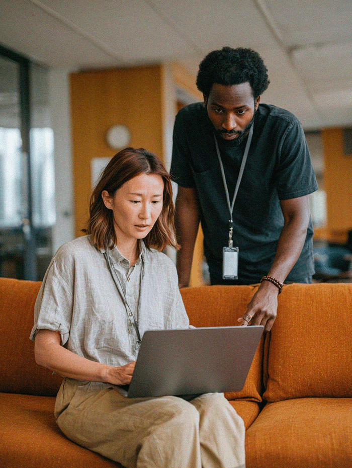 A woman sits on an orange couch using a laptop, focused on the screen. A man wearing a lanyard stands beside her, leaning slightly forward and pointing toward the laptop as if explaining or assisting with something. The setting appears to be a modern office or collaborative workspace with warm lighting and glass-walled rooms in the background.