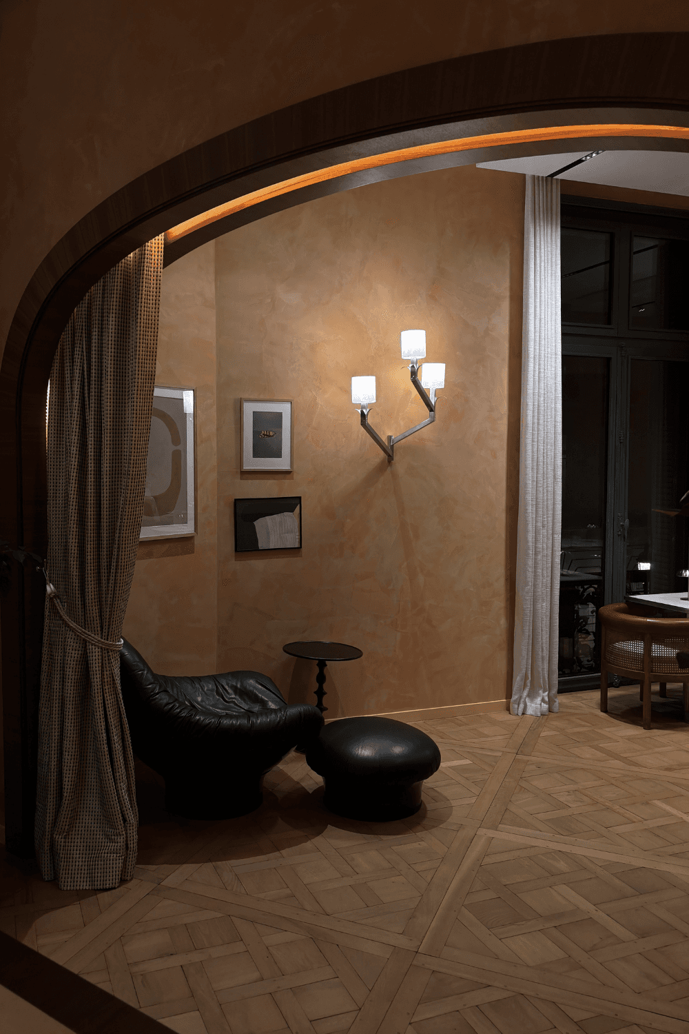 A woman in black stands focused over a laptop on a table, surrounded by colorful paper samples pinned to a white wall, creating an artistic, thoughtful atmosphere.