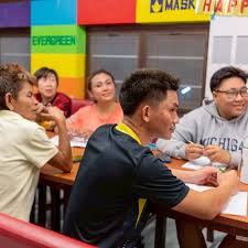 man in blue t-shirt and white floral skirt holding black smartphone while sitting on sidewalk