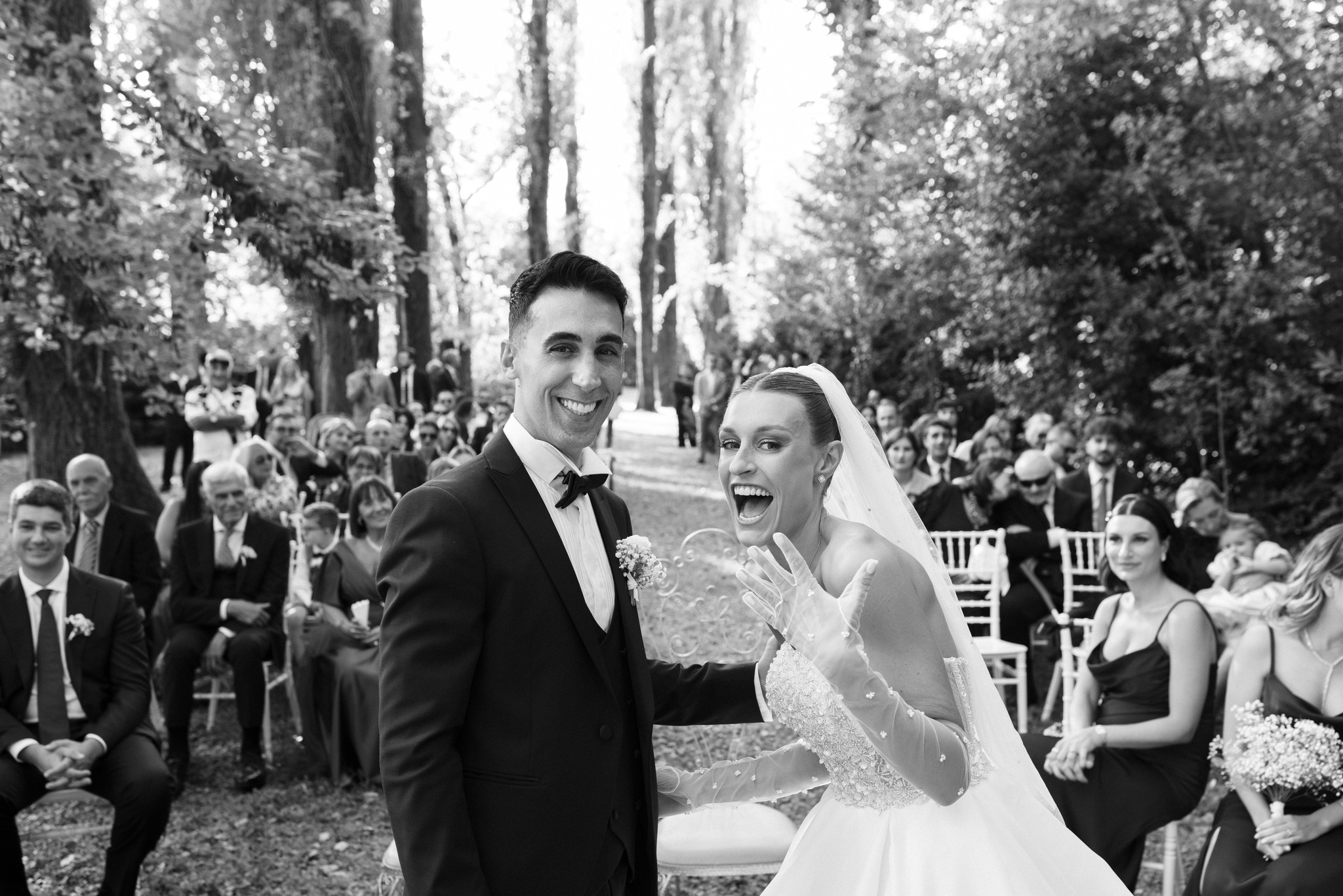 Eleonora e Fabio si sorridono durante la cerimonia nel parco di Villa Ferrari, tra gli ospiti e i cipressi