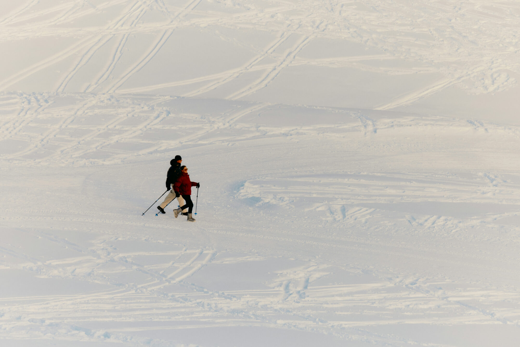 Two people walk across a snowy landscape with ski poles, leaving trails in the fresh snow. The scene conveys calm and solitude.