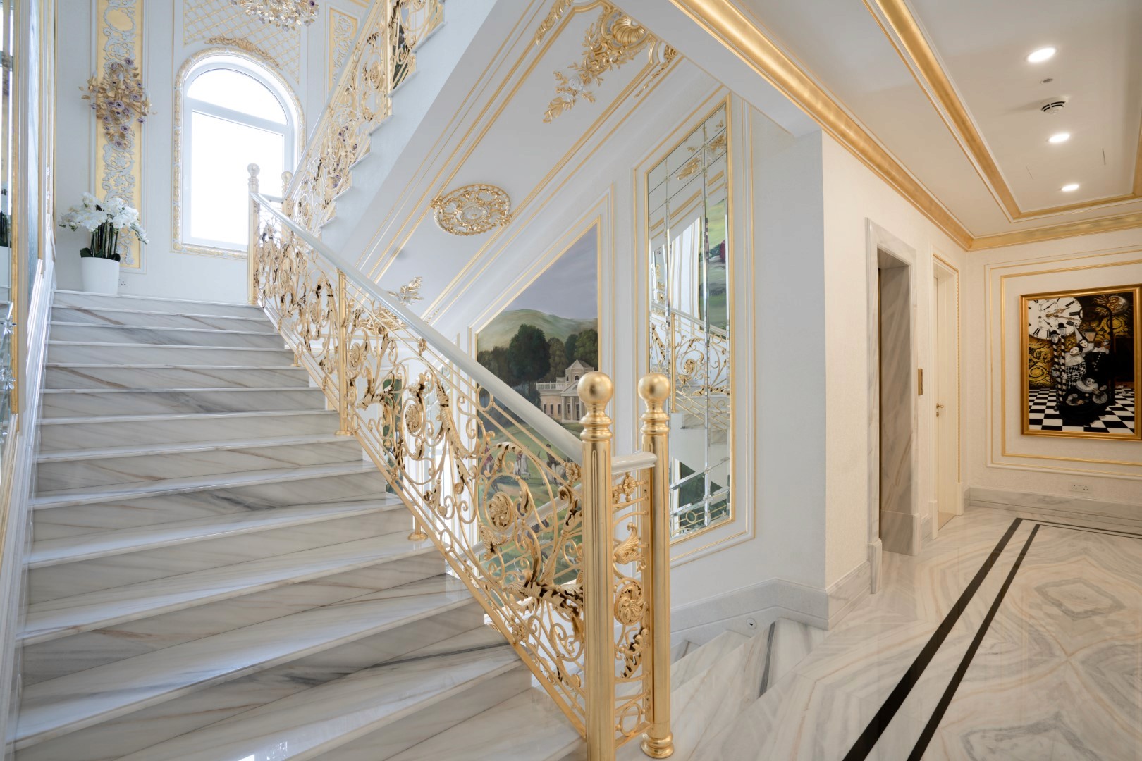 Luxurious marble staircase with ornate golden railings leads to a brightly lit hallway adorned with elegant artwork and intricate moldings, showcasing opulent interior design.